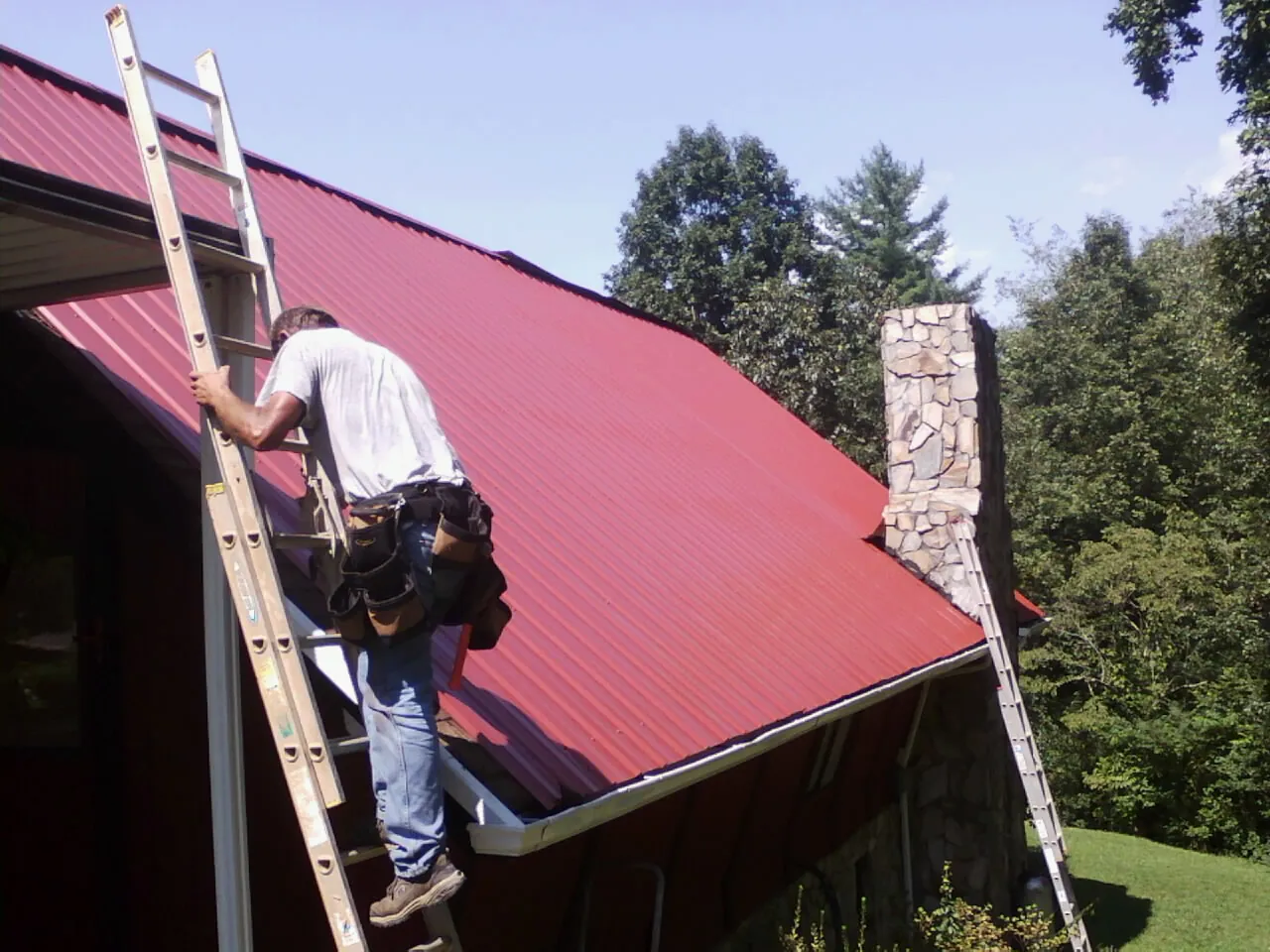 Roofer on a ladder beside a red metal roof and stone chimney — Soffit & Fascia Repair by Roofing Brothers in Zuni